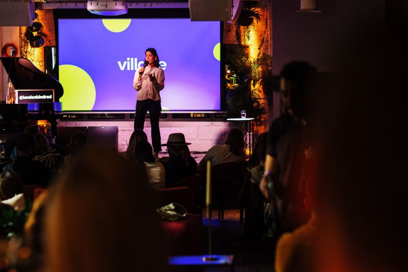 Jules speaking on stage at Kindred London's ID8, with a Village presentation behind her and the audience in the foreground.